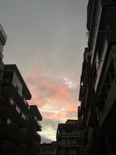 The image displays an urban residential scene in Karşıyaka, Turkey, observed at dusk or dawn. The central focus is a cloudy sky, exhibiting a palette of orange, pink, and white hues, indicative of low-angle sunlight, transitioning to a dull grey in the upper portion. Flanking this sky are two multi-story residential buildings. The building on the left, partially visible, is lighter in color and features multiple balconies, some enclosed with glass panels and others with metal railings. An external air conditioning unit is mounted beneath one balcony. The building on the right is largely silhouetted against the illuminated sky, appearing darker. It also presents multiple balconies and windows, with some visible external window coverings. A small cluster of green foliage is evident near the top edge of the right building. The perspective is upward, suggesting the photograph was taken from a street or alleyway situated between these structures. No people, vehicles, or street-level activities are discernible.