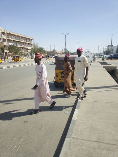 The image displays a sunlit street scene in Gyadi Gyadi Arewa, Nigeria. Three adult males are walking from left to right across the foreground. The individual on the left wears a light pink traditional robe and a red and white striped cap. The central figure wears a brown traditional robe and a similar red and white striped cap; this individual is partially obstructed by a yellow tricycle (Keke Napep) positioned directly behind them, facing away from the camera. The third individual, on the right, wears a cream-colored traditional robe, a red baseball cap, and sandals over socks, with a slight smile directed towards the camera.
The environment features a paved asphalt road with a black and white striped median barrier running into the distance. A multi-story brown building with multiple windows stands on the left side of the road, behind several palm trees. Another yellow tricycle is visible further down the road on the left, along with other vehicles and parked cars. On the right, a concrete sidewalk is present, with debris including a plastic bottle and string. Beyond the sidewalk, more parked cars, utility poles, and streetlights with solar panels are visible. A mosque minaret is discernible in the distant background. The sky is clear and blue, and prominent shadows are cast to the right, indicating bright sunlight.