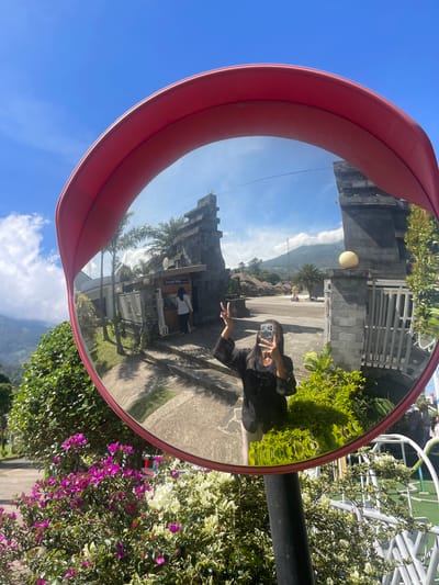 A person is reflected in a convex traffic mirror along a roadside in Kledung, Indonesia. The mirror's reflection shows a paved area, a building with stone walls and a gate, a distant mountain, and a cloudy sky. The person is holding a phone, seemingly taking a picture, and making a peace sign. The mirror itself has a red frame and is mounted on a black pole. Lush greenery, including flowering bushes with purple blooms, surrounds the pole.
