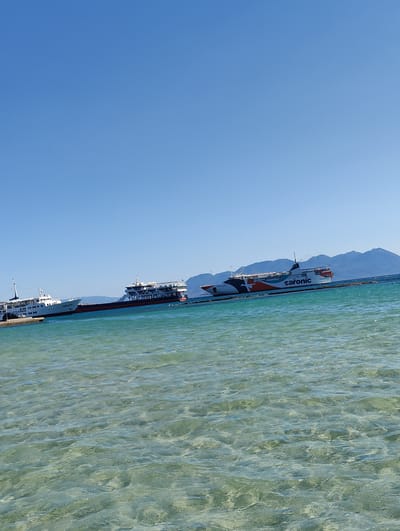 The image shows a clear blue sky over a calm, turquoise sea. In the distance, mountains are visible. The foreground consists of shallow, clear water with visible ripples. Several large ferries are docked or cruising in the mid-ground. One ferry, labeled "SARONIC," is prominent, with orange and white markings. Another ferry, white with "COSMOS" written on its side, is docked closer to the shore. A third ferry, with a red hull, is visible behind the "COSMOS" ferry. The scene appears to be a port or harbor area, likely in or near Aegina, Greece, given the presence of the "Saronic" ferry line, which operates in the Saronic Gulf. There are no people clearly visible in the image.