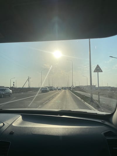 The image is taken from the interior of a vehicle, looking forward through the windshield. The sun is bright and low in the sky, causing lens flare and washing out the upper portion of the image. The road ahead is a two-lane highway with streetlights lining both sides. Several cars are visible on the road, driving in the same direction as the camera. To the right of the road, there is a pedestrian walkway and some construction material. A triangular road sign with a symbol of a truck and a speed limit of 70 is visible on the right side of the road. In the distance, there are some low-rise buildings and utility poles. The scene appears to be in a semi-urban or suburban area, possibly on the outskirts of a city. The specific location is Баянды ауылдық округі, Kazakhstan.