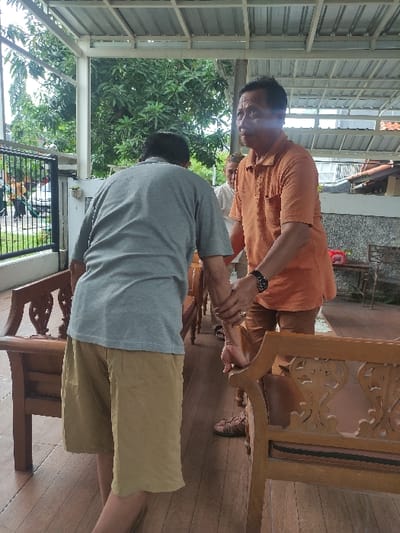 The image, taken in Lamongan, Indonesia, captures a daytime scene featuring two men in a covered outdoor or semi-outdoor setting. The photo is rotated 90 degrees counter-clockwise.
In the foreground, a man wearing an orange short-sleeved collared shirt and a dark wristwatch is visible from the chest up, facing towards the viewer. He has dark hair and glasses, and his expression is focused. His right hand is clasped around the wrist or hand of another man.
This second man is positioned with his back mostly towards the viewer, wearing a grey t-shirt and khaki-colored shorts. He is leaning or bending forward, and his left arm is extended towards the first man's hand.
Behind the two men, lush green foliage, likely trees or bushes, is visible, suggesting a natural outdoor environment. A structure with a white corrugated metal roof and supporting beams frames the top and left side of the scene. On the right side of the frame, a ornate, light-colored wooden table or bench with carved details is present. Further in the background, a bird cage with a bird inside can be seen near the ground. The floor appears to be made of wooden planks.
The overall atmosphere suggests a casual, possibly informal gathering or interaction. There is no discernible text visible in the image.