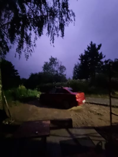 The image depicts an outdoor scene at dusk, with a purplish-blue sky suggesting twilight or a storm. In the foreground, a dark, out-of-focus area suggests a seating area with wooden furniture. A red convertible car, appearing wet, is parked on a dirt or gravel surface in the middle ground. Beyond the car, a dense line of trees forms the background. The environment appears to be rural or semi-rural. The location is identified as Szczaki, Poland, though no specific landmarks within the city are visible.