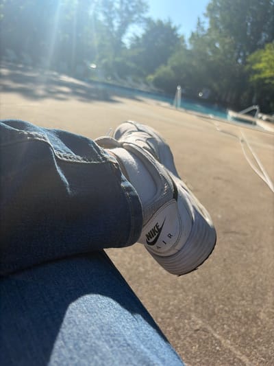 A close-up, angled shot focuses on a person's left leg clad in blue jeans and a white Nike Air sneaker, resting on a concrete surface, presumably outdoors. The jeans are denim with visible stitching and a pocket hem. The shoe exhibits the Nike logo. The background is blurred, showing trees and a sunlit area, possibly near a body of water. While the general location seems to be outdoors in a place with trees and concrete, there is insufficient data to determine the specific location in Edison, United States of America.