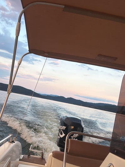 The image depicts a view from the stern of a boat moving through calm water, likely a lake or a wide river, in the vicinity of Zvanika, Bulgaria. The water is disturbed by the boat's wake, creating white foam. A dark outboard motor, branded "MERCURY," is visible towards the right of the frame. The boat appears to have tan upholstery and a tan canopy. In the distance, a tree-covered shoreline with rolling hills is visible under a sky filled with soft clouds tinted with the colors of dusk, suggesting late evening. The overall impression is one of peaceful travel on the water at sunset. There are no people visible in the frame.