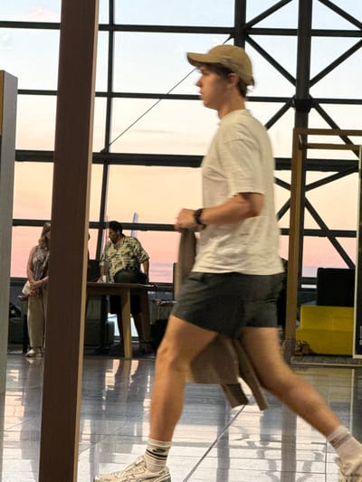 In the foreground, a young Caucasian male wearing a light-colored t-shirt, dark shorts, a beige baseball cap, and white athletic socks with black stripes on the ankle, is walking. He is wearing white sneakers and a black watch. His right leg is extended forward as he moves. The background is blurred, but it appears to be an indoor setting with large windows showing a sunset.
Behind the man, a woman with blonde hair, wearing a patterned sleeveless top and beige pants, stands to the left. To her right, a man with dark hair and a beard, wearing a short-sleeved patterned shirt and dark shorts, is standing behind a wooden table. A black bag rests on the table. The setting appears to be an airport terminal, as evidenced by the large windows, metal framework, and reflective floor. The photograph was taken in the city of Zaventem, Belgium, likely at Brussels Airport.