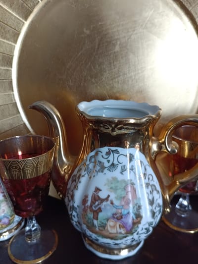 The image displays a close-up view of several antique porcelain items arranged on a dark surface, with a large, textured golden plate serving as a backdrop. In the foreground, a stemmed glass with a ruby-red interior and intricate gold patterns is positioned to the left. To its right, a golden teapot with a long, curved spout is prominently featured. Adjacent to the teapot, a small porcelain jug or creamer showcases a painted scene of a man playing a lute and a woman holding a basket of flowers, all adorned with gold trim. A second, partially visible golden teapot or pitcher is seen on the far right. The objects appear to be decorative and are likely part of a tea set. The overall lighting is warm, highlighting the metallic sheen of the porcelain. The location in Évreux, France is not evident from the image itself.