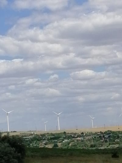 The image displays a landscape featuring several wind turbines against a backdrop of a cloudy sky. The turbines, with their white towers and blades, are scattered across the scene, some in the mid-ground and others further in the distance. They are positioned above a town or village, characterized by numerous houses with red roofs and green vegetation, nestled on a hillside. The foreground shows a field of green and brown grass.
The sky is filled with a mix of blue patches and large, fluffy white and gray clouds, suggesting a partly cloudy day. The lighting indicates it is daytime, likely late morning or afternoon, given the brightness. The scene appears to be a rural or semi-rural setting, possibly a wind farm located near a residential area. There is no visible text in the image. The overall mood is calm and expansive, highlighting a blend of natural scenery and renewable energy infrastructure. The location context provided is Cernavoda, Romania.