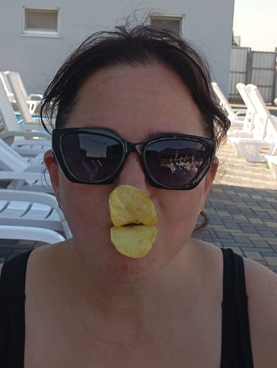 The media file shows a close-up shot of a woman in an outdoor setting, possibly a resort or pool area, given the presence of lounge chairs and paved ground. The woman is wearing large, black sunglasses, which reflect a scene with more lounge chairs and a building. Two potato chips are placed on her lips, giving the appearance of a playful or humorous expression. Her hair is dark and styled loosely. The background suggests a sunny day, and the overall mood appears lighthearted and casual. The woman is wearing a black tank top. The location is identified as Vladimirovka, Moldova. No visible text is present in the image.