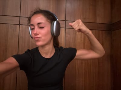 A woman hangs upside down, wearing headphones and a black t-shirt. Her eyes are closed. She is in a wood paneled sauna. Her arms are outstretched, one with a clenched fist. The background consists of vertical wood panels.