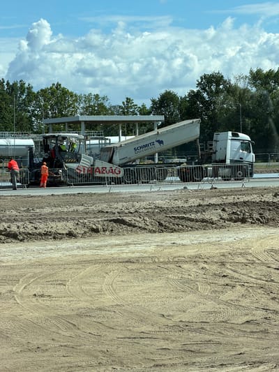 In Krzeptów, Poland, on a construction site, a road paving operation is underway. A paving machine, painted white with red lettering that reads "STRABAG", is actively laying down asphalt. A large white dump truck, identified as "SCHMITZ CARGOBULL", is positioned behind the paver, its bed tilted upwards, discharging material. Two construction workers are present: one in an orange high-visibility suit stands near the rear of the paver, and another in an orange vest and pants is on the left side of the paver, observing the process. A metal barricade separates the active work area from the surrounding ground. The environment is characterized by a partially constructed road surface, with areas of loose soil and tire tracks indicating recent activity. In the background, trees and a covered structure are visible under a bright blue sky with scattered white clouds.