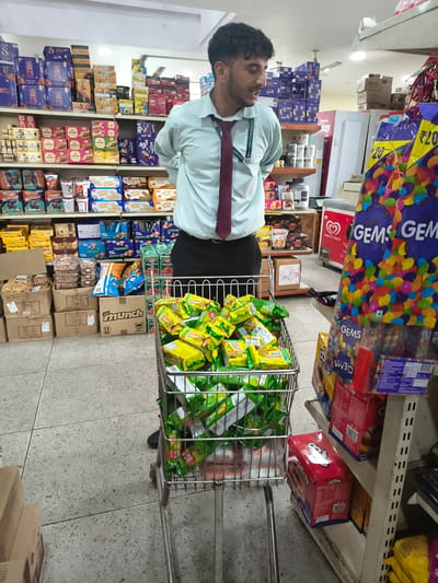 In a retail store in Sidhpur, India, a young male employee wearing a light blue shirt and a maroon tie stands near a shopping cart overflowing with green-packaged snacks. The cart's contents appear to be individual snack portions, possibly biscuits or wafers, with some portions of larger snack bags also visible at the bottom. Behind the employee, shelves are stocked with various consumer goods, including boxes of chocolates, packaged snacks, and other food items. A large display of colorful candies in transparent packaging is situated to the right of the employee. The store has a tiled floor and fluorescent lighting, typical of a supermarket or convenience store environment. The employee is looking towards the shopping cart and appears to be overseeing the stocked items.