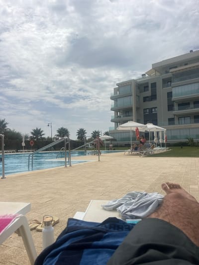 The image depicts a daytime scene at a swimming pool area in Ayamonte, Spain, under an overcast sky. In the immediate foreground, the viewer's lower legs and feet are visible, covered partially by a black garment. A blue and black striped towel lies on a light-colored lounge chair, next to which a white water bottle and light-colored flip-flops are positioned. Another grey towel is on a second lounge chair to the right.
The midground features a large, rectangular swimming pool with light blue water, accessible by multiple stainless steel handrails. The pool is surrounded by a light-colored tiled deck. Beyond the pool, a young child in a light-colored swimsuit stands on the deck. Further back, on a patch of green grass, several white sun loungers are arranged under two white sun umbrellas. Multiple individuals, including adults and children, are seated or reclining on these loungers. A red lifebuoy is attached to a pole near one of the umbrellas.
In the background, a modern, multi-story building with a light-colored facade and numerous glass balconies dominates the right side of the frame. Palm trees are visible along the distant horizon, particularly on the left side, behind a low fence or hedge. The sky is largely covered with patchy white and grey clouds.
