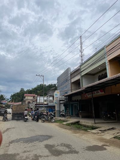 The image depicts a street scene in the city of Rantau Pauh, Indonesia, under a cloudy sky. A two-lane asphalt road runs through the frame, with a truck and several motorcycles present. The truck, laden with goods, is positioned on the left side of the road, emitting exhaust smoke. A motorcyclist is visible on the left, and several other motorcycles are parked or being ridden by individuals on the right side of the road.

Along the right side of the street, a row of buildings lines the road. These buildings appear to be commercial establishments, with awnings and signs indicating their purpose. One sign reads "BSI BANK" and "ATM", and another displays "APOTEK 3R". The buildings have multiple stories and are constructed with various materials, including concrete and colored siding.

On the left side of the road, a smaller "MINI MARKET" sign is visible. Beyond the immediate storefronts, more buildings and lush green foliage, including palm trees, are present on a rising hillside. A cellular tower is also visible in the distance. The overall environment suggests a moderately developed urban or semi-urban area.