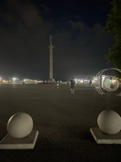 At night, a wide paved plaza is depicted, with a tall column topped by a statue in the background. In the foreground, two large spherical white objects on square bases flank the view. Further into the plaza, a man and a child are walking away from the camera, and several modern artistic installations resembling large hoops are visible to the right. Streetlights line the horizon in the distance, illuminating the scene. The location is identified as the city of Berezhnoi, Russia.
