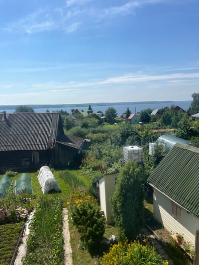 The image depicts a rural landscape under a clear blue sky with wispy clouds. The foreground shows a garden with rows of vegetables and flowers, including some covered with protective netting or plastic sheeting. A path runs through the garden, bordered by greenery and flowering plants.
In the middle ground, several structures are visible. To the left, a weathered, dark-colored wooden building with a corrugated roof dominates the scene. Adjacent to it, there are more garden structures and a large white water tank. Further into the scene, a greenhouse or hoop house is present, along with other smaller buildings and trees. A second building with a green corrugated roof is situated to the right, with a light-colored wall and a wooden bench outside.
In the background, a body of water, likely a river or lake, stretches across the horizon. Several houses with green and red roofs are scattered along the shoreline, interspersed with trees. A tall green spire, possibly belonging to a church, is visible in the distance. The overall impression is that of a peaceful, summer day in a village or rural settlement, possibly Gari, Russia, given the architectural style and natural setting. There are no people visible in the image.