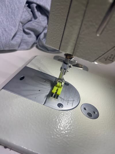 The image shows a close-up view of a sewing machine needle and presser foot. A piece of grey fabric is draped over the top left of the sewing machine. The presser foot is yellow and is holding the fabric in place. A black thread is visible, leading to the needle. The sewing machine is white and appears to be made of metal and plastic. There are measurement markings on the sewing machine's plate. The scene suggests an indoor, domestic or workshop setting, with a focus on the mechanics of sewing. The provided location of Novogeorgiyevka, Russia, is not directly observable or inferable from the visual information within the image itself.