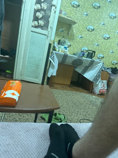 The image depicts an indoor scene, possibly a room in a residential dwelling in Corlăteni, Moldova. The room is decorated with patterned wallpaper and appears somewhat cluttered. In the foreground, a person's legs, clad in black socks, are visible, resting on a pink surface, likely a bed or sofa. To the left, an orange rectangular object, possibly a food package, rests on a brown table. Behind the table, a doorway with a patterned glass panel is visible, leading to another part of the dwelling. In the background, a table is covered with a white tablecloth adorned with a Greek key pattern and a circular motif. On this table, several bottles of water and a black electric kettle are present. Further in the background, a white plastic bag with a red logo that reads "Kaufland" and "Jeftin" is visible. Green rubber slippers, shaped like cartoon characters, are placed on the floor near the table. The overall impression is of a lived-in, domestic environment.