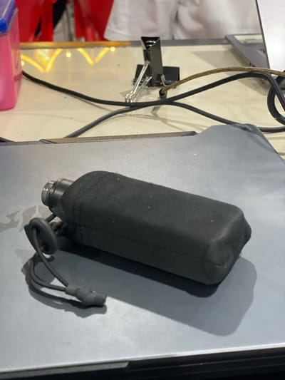 The image depicts an indoor scene in Lhok Mereubo, Indonesia, primarily focusing on a desktop setup. In the foreground, a black, rectangular object, possibly a flask or power bank, is horizontally positioned on a closed grey laptop or tablet lid. This object is covered in a dark, textured material and features a silver-colored metallic top with an attached black drawstring and stopper on its left end.

Behind the laptop/tablet, a light-colored tabletop is visible, cluttered with multiple black electrical cables and one brown cable. A black binder clip stands upright on the tabletop, positioned amidst the cables. To the far left of the tabletop, a portion of a pink plastic container is discernible. In the blurred background, red objects, likely seating or decorative elements, are present, along with yellow string lights. A person wearing a white shirt is partially visible behind the table. No active interactions or specific actions are depicted; all subjects and objects are stationary.