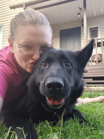 A young woman with blonde hair pulled back and wearing round glasses and a pink t-shirt is leaning into a black dog. She is making a kissing gesture towards the dog, whose head is turned towards the camera. The dog has brown eyes and its mouth is slightly open, revealing its tongue. They are both on a grassy lawn in front of a house with a light-colored siding and a porch. The dog's fur is dark and appears to be short to medium length. The woman's skin is fair. The background shows a dark gray door and a window on the house. There are also railings and furniture visible on the porch. The scene appears to be outdoors during the daytime. The location is the city of Koledino, Russia.
