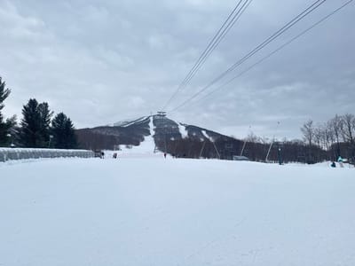 A snowy landscape under an overcast sky features a prominent mountain in the distance, its slopes marked by cleared paths. A ski lift structure sits atop the peak. Skiing infrastructure, including chairlifts and fencing, is visible mid-ground. A snow-covered field occupies the foreground, with a low structure on the left and scattered trees throughout the scene.
