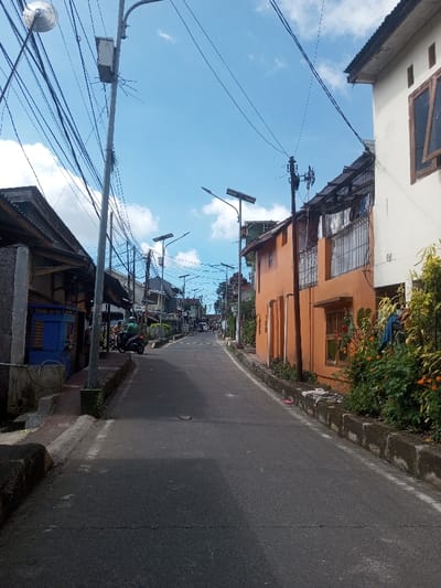 The image displays a street scene in Naung Bo Oe, Myanmar, under a clear blue sky with scattered white clouds. A two-lane asphalt road, sloping gently uphill and curving slightly to the left, occupies the center. Concrete curbs delineate the road from the adjacent structures and sidewalks.

On the left, a single-story building with a dark roof and a blue-painted wooden structure is visible. A tall, grey utility pole stands prominently in the foreground, equipped with a traditional street lamp fixture, a grey utility box, and a dense bundle of black electrical wires extending overhead. Further down the street, a black scooter is parked on the sidewalk near two individuals, one seated on the scooter and one standing. Several more utility poles, each featuring a solar panel and LED streetlight, line the left side of the street.

On the right, a two-story building with an orange-painted exterior and dark-framed windows is present, followed by a section of a lighter-colored building with similar windows. A concrete curb separates the road from a raised planter containing green shrubs and orange-flowering plants; a blue garment is draped over some of the foliage. Additional overhead wires are strung along the right side, connected to multiple utility poles, some with solar-powered streetlights. Sunlight casts distinct shadows on the road surface and buildings.