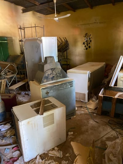The image shows the interior of a room in Khartoum, Sudan, located at coordinates 15.552 latitude and 32.57 longitude. The room is cluttered with old appliances, including a refrigerator, a freezer, a television set, and a window air conditioner. The appliances are dirty and covered in dust. The room is also filled with debris, such as cardboard boxes, pieces of wood, and electrical cords. There are no people present. The room is dimly lit. The ceiling is made of wood and has a fan hanging from it. The walls are yellow and have a floral design painted on them. The floor is made of dirt and is covered in debris.