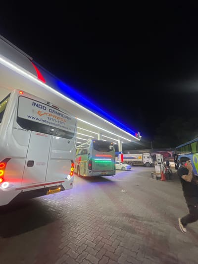 The image depicts a nighttime scene at a gas station in Kajheri, India. The foreground features the rear of a white bus with "INDO CANADIAN EXPRESS" branding and contact information. To the right of this bus, another bus is visible, also with illuminated signage. In the middle ground, a white car is parked, and a truck with "HP Power95" branding is also present. The gas station features blue and white illuminated structures overhead. In the background, several other vehicles and what appear to be fuel pumps are situated. A person dressed in dark clothing is on the right side of the image, talking on a mobile phone. The ground is paved with interlocking bricks, and shadows are cast across the scene.