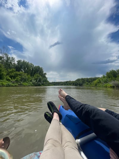 The image captures a serene scene on a river, likely the Irtysh River near the village of Samorodovo, Russia, given the characteristic landscape. Two individuals are relaxing in what appears to be an inflatable raft or kayak, their legs extended towards the camera. One person is wearing khaki-colored corduroy pants and a sandal on their left foot, while the other is clad in dark pants and black water shoes with green accents.
The raft, predominantly blue, rests on the calm, greenish-brown water of the river, which reflects the overcast sky and the lush, green foliage lining its banks. The riverbanks are densely wooded with deciduous trees, creating a natural, unspoiled vista. The sky is dramatic, with a mix of dark, heavy clouds and patches of brighter sky, hinting at changeable weather. Several small bubbles are visible on the water's surface, suggesting gentle movement or aeration. The overall atmosphere is peaceful and suggests a leisurely activity enjoyed in nature.