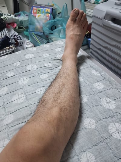 A person's bare leg and foot are visible, resting on a gray blanket with a white floral pattern. The leg is covered in hair, and the foot shows toenails. In the background, various items are piled up, including plastic bags, clothing, and what appears to be a cooler. The setting seems to be indoors, possibly a room in Anseong-si, South Korea, given the context provided.