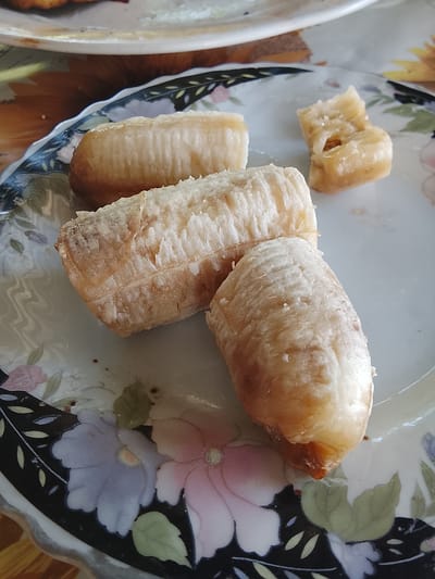 The image shows a close-up of a white plate with a floral pattern, containing four pieces of peeled, cooked bananas. Three of the pieces are whole and oblong, while the fourth is a smaller, broken fragment. The bananas appear slightly softened and have a light brown hue in some areas, suggesting they may have been fried or baked. The plate has a decorative border with black and colorful floral designs. The setting appears to be indoors, possibly a kitchen or dining area, with a patterned tablecloth visible beneath the plate. The location is identified as Жалпақсай, Kazakhstan. There are no people or animals visible in the image, and no specific actions are depicted beyond the arrangement of the food on the plate.