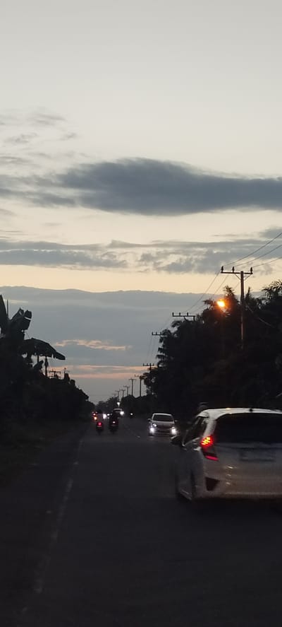 The image depicts a street scene in Kruengseupeng, Indonesia at dusk or dawn. The road extends into the distance, flanked by trees and utility poles. Multiple vehicles, including cars and motorbikes, are visible on the road. The sky is overcast with layered clouds. A street light is illuminated on the right side of the road.