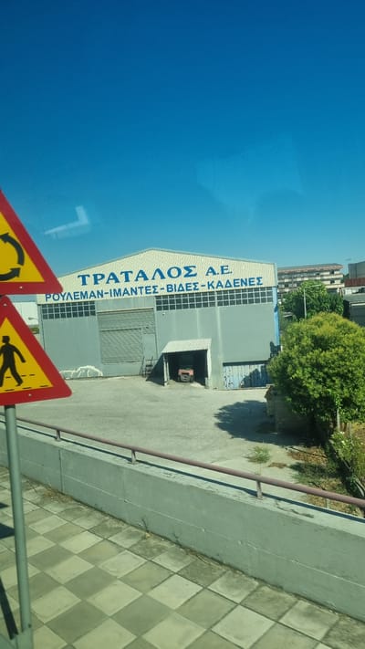 The image captures a wide shot of a commercial building with "ΤΡΑΤΑΛΟΣ Α.Ε." and "ΡΟΥΛΕΜΑΝ-ΙΜΑΝΤΕΣ-ΒΙΔΕΣ-ΚΑΔΕΝΕΣ" written in blue capital letters on its upper facade. The building appears to be a warehouse or factory, with a large, grey, corrugated metal roll-up door in the center. To the right of the main entrance, there's a smaller, open-fronted structure housing a red piece of machinery, possibly a forklift. The building's exterior is grey, with a row of windows above the main entrance and a light-colored, ribbed roof.
In the foreground, two triangular yellow and red traffic signs are visible on a metal pole. The upper sign displays a circular arrow graphic, while the lower sign shows a silhouette of a person walking. A low, concrete barrier with a reddish-brown railing runs horizontally across the lower portion of the image, separating the building's lot from a checkered tiled walkway.
The environment suggests a sunny day, with a clear blue sky dominating the upper part of the frame. A dense green tree is situated to the right of the building, casting a shadow on the ground. In the background, other buildings and trees are visible, indicating an urban or industrial area. The image appears to have been taken from a moving vehicle, possibly a bus or car, due to the slight distortion and reflection on the window. The presence of the Greek text strongly suggests the location is in Greece, and the specific company name and product offerings (bearings, belts, screws, chains) are indicative of an industrial or commercial establishment. Based on these cues, the location is likely within the city of Larissa, Greece, known for its industrial activity.