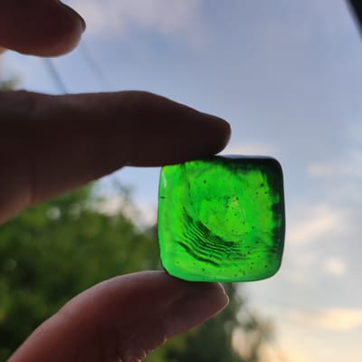 A close-up, eye-level shot captures a person's thumb and index finger holding a small, translucent, green, cube-shaped object. The object is positioned in the center of the frame, with the fingers creating a partial frame around it. The background is out of focus, showing a blurred green foliage, suggesting an outdoor environment, possibly a park or garden. Above the foliage, the sky is visible, with soft clouds and a hint of sunset light. The overall lighting is gentle, with the sun likely setting, casting a warm glow. There are no visible signs or indications suggesting the location is specifically in Kineshma, Russia.