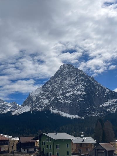 The image captures a view in the city of Sappada, Italy. In the foreground, there are buildings, one of which is green, suggesting an alpine village setting. The central focus is a large, snow-capped mountain, presumably part of the Dolomites, against a partly cloudy sky. The mountain's slopes are steep and textured, with visible rocky areas. The mountain is covered with a dense, evergreen forest that appears to ascend its lower slopes. The sky features scattered clouds and areas of clear blue, indicating a fair-weather day.