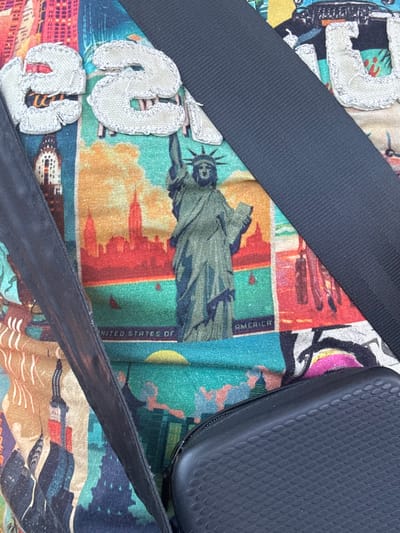 The image shows a close-up of a colorful t-shirt with a collage of New York City landmarks. The Statue of Liberty is prominently displayed in the center, along with images of the Manhattan skyline and possibly other iconic buildings. The text "UNITED STATES OF AMERICA" is visible below the Statue of Liberty. The t-shirt has appliqued letters that spell out "USA." A black strap, possibly from a bag or seatbelt, diagonally crosses the t-shirt. In the lower right corner, a black zippered pouch with a textured surface is visible. The overall impression is that of a tourist souvenir t-shirt. There is no indication of the city of Sidi H'Mad Ou M'Barek, Morocco, in the image.