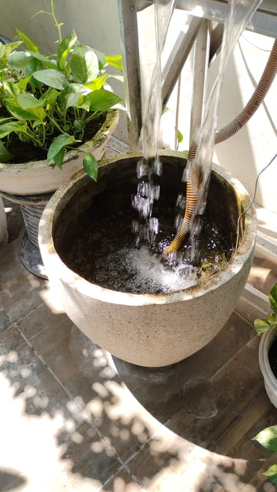 In Banjarsari, Indonesia, a close-up shot captures a decorative outdoor fountain. Water cascades from an unseen source into a large, speckled, light-colored basin. A flexible, corrugated brown tube is partially submerged in the basin, likely part of the water circulation system. To the left, a potted plant with large, variegated green leaves sits beside the basin. The ground beneath is tiled with a pattern of brown and gray squares. Sunlight streams from the upper right, casting dappled shadows of leaves and branches onto the tiles, suggesting the presence of overhead foliage. The scene appears to be on a patio or balcony.