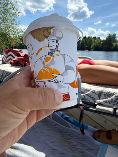 The image shows a hand holding a paper cup with a cartoon baker character on it. The cup is partially filled with a pastry. In the background, a person in a red bikini is lying on a lounge chair on a wooden dock by a body of water. Trees and a blue sky with clouds are visible. The scene appears to be outdoors on a sunny day, likely in the city of Dobryanka, Russia, given the presence of a lake and lush vegetation.