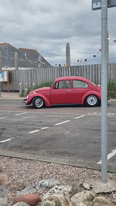 This media file shows a red classic Volkswagen Beetle parked on a street. The car is the primary subject, viewed from its side. It has chrome detailing on the bumpers and door handles, and classic silver alloy wheels. The driver, wearing a hat, is visible through the tinted windows.

The setting appears to be a coastal town, possibly Montrose, United Kingdom. In the background, there is a wooden fence, a building with a grey slate roof adorned with colorful bunting, and some vegetation. The street is paved with asphalt and has white lane markings. To the right of the street, there is a curb with an area of small pebbles and larger, smooth stones, some of which are speckled.

The sky is overcast, suggesting a cloudy day. The overall mood is relaxed and perhaps a bit nostalgic, given the vintage car. There is a signpost visible near the front of the car, but the text is not legible.