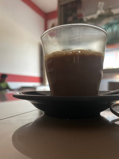 A close-up, low-angle shot captures a glass of dark brown liquid, likely coffee or tea, sitting on a black saucer. The liquid has a foamy top layer. The background is blurred, showing a white wall with a red stripe and a hint of a window with green foliage outside. In the distance, a person wearing a green shirt is out of focus. The scene appears to be inside a cafe or restaurant in Cotseutui, Indonesia. The lighting is dim, creating a soft shadow of the glass and saucer on the table.
