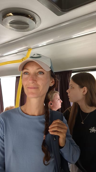 In Malyi Mayak, Ukraine, three individuals are observed inside a public transportation vehicle. In the foreground, a woman wearing a light blue sweatshirt and a baseball cap with a smiling face design is looking away from the camera. Her brown hair is braided and hangs over her shoulder. She is holding a dark blue bag with a visible Nike logo. To her right, a young woman with long brown hair and a black t-shirt is also looking away. Behind the woman in the foreground and to her left, another woman with blonde hair and earbuds is partially visible. The interior of the vehicle features a yellow handrail and a circular air vent on the ceiling, consistent with a bus or similar public transport. Brown curtains are visible in the background.