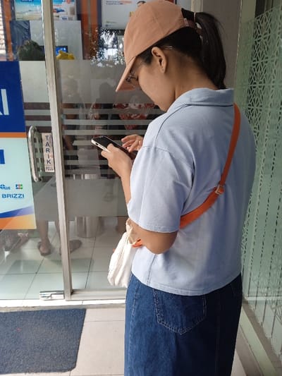 A person is depicted from behind, standing outdoors near a glass-fronted entrance in Sarimulyo, Indonesia. The individual wears a light peach baseball cap, clear-rimmed glasses, a light blue short-sleeved collared shirt, and a long dark blue denim skirt. Their dark hair is pulled back into a ponytail. Both hands are actively engaged with a black smartphone, suggesting interaction with its screen. An orange strap from a shoulder bag crosses their back, with a light-colored bag visible on their right hip.
To the left, a glass door features a silver pull handle with vertically printed Indonesian text "TARIK" and English text "PULL." Below these labels, "BRI," "BRIZZI," and "BRIZZI PLUS" logos are present, indicating a Bank Rakyat Indonesia (BRI) facility. Reflections on the glass door show the lower legs and feet of individuals inside, some wearing sandals, and a metallic cylindrical object. Through the glass, distorted interior details, including advertisements, are partially visible. The ground is a light-colored tiled surface, with a dark blue rectangular doormat positioned in the lower-left foreground. To the right of the subject, a patterned glass panel with a repetitive geometric design forms part of the building's facade.
