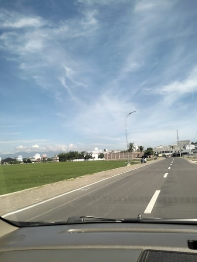 The image depicts a view from inside a vehicle traveling along a paved road. To the left of the road is a vibrant green rice paddy field, which extends towards a row of buildings and trees in the distance. The buildings are a mix of residential and commercial structures, with some featuring white domes and facades, suggesting a religious or institutional presence. A tall, modern lamppost stands beside the road, and further down, a cell tower is visible against the blue sky. Several motorcycles are parked or in motion on the side of the road. The sky is partly cloudy with wispy white clouds scattered across a clear blue expanse. The scene is set in Keude Lueng Putu, Indonesia, characterized by its agricultural landscape and developing urban elements.