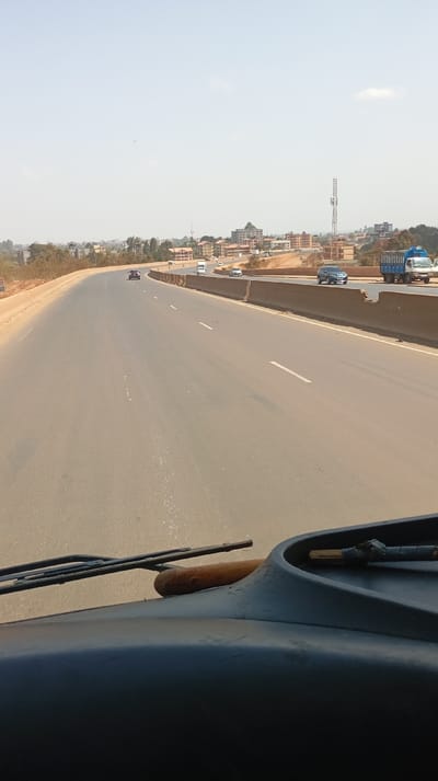 The image depicts a multi-lane highway scene in Gitaru ward, Kenya, viewed from the interior of a vehicle. The foreground shows the vehicle's dark dashboard and a windshield wiper resting on the left side. The asphalt highway stretches into the distance, appearing dry and wide with visible white dashed lane markings. To the right, a long concrete barrier separates the main thoroughfare from an adjacent paved area, possibly a service road or exit ramp.
Multiple vehicles are present on the highway. In the mid-ground, a dark-colored SUV is observed in the leftmost lane. Further down, a light-colored box truck, a white semi-truck, a white van, and several passenger cars are visible. On the paved area to the right of the concrete barrier, a blue and white box truck is positioned. No people are discernible.
The surrounding environment consists of dry, brownish vegetation lining the left side of the highway. In the background, an urban landscape is visible with numerous buildings of varying sizes, predominantly featuring reddish-brown roofs, situated on elevated terrain. A prominent, tall lattice telecommunication tower stands amidst these buildings on the right side of the frame. The sky is largely clear and pale blue, indicating a sunny day, with a small, wispy white cloud present in the upper right quadrant.