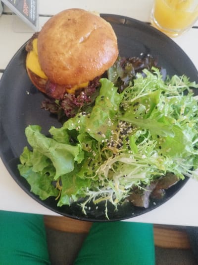 A top-down shot reveals a black plate featuring a burger, accompanied by a vibrant green salad. The burger boasts a glossy bun, cheese, and bacon peeking out. The salad is dressed with a glistening sauce and sprinkled with black and white sesame seeds. A glass of orange juice sits partially visible near the plate's edge. A glimpse of someone wearing green pants is also present in the image.