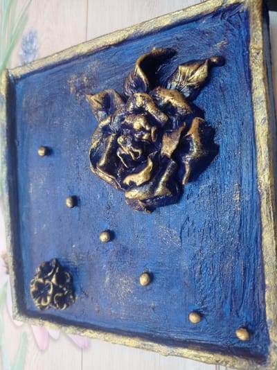 A close-up, angled shot captures a textured, rectangular artwork mounted on a light-colored, wood-grain surface. The artwork itself is predominantly a deep, textured blue with prominent gold accents. A large, three-dimensional sculpted rose, painted in a dark, metallic blue with highlights of gold, dominates the upper portion of the artwork. Its petals are intricately detailed and appear to be made of a malleable material. Scattered across the blue surface are several small, raised gold spheres, resembling beads or dots. In the lower left corner, a smaller, less distinct sculpted flower, also in gold and dark blue, is present. The edges of the artwork are framed with a thick band of gold paint. The background surface suggests an indoor setting, possibly a wall or shelf, with a hint of out-of-focus floral wallpaper or a decorative item visible in the far left and lower edges of the frame. The artwork appears to be a decorative piece, possibly intended for wall display. The location, Дукорскі сельскі Савет, Belarus, is not visually represented in the image itself.