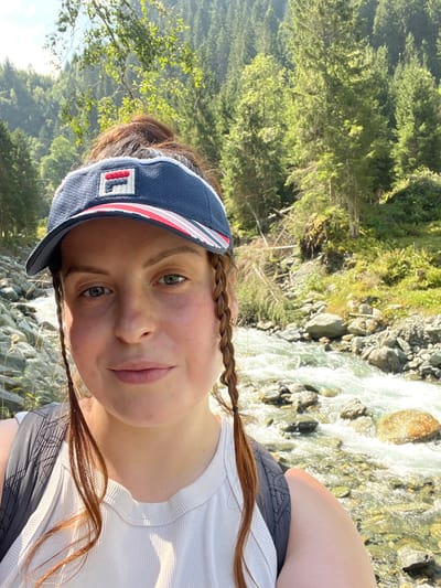 A close-up selfie depicts a single female subject in an outdoor, mountainous environment consistent with the region of Reitlehen, Austria. The subject, positioned centrally, has light skin, blue eyes, and reddish-brown hair styled in a high bun, with two thin braids framing her face. She wears a navy blue Fila visor with red and white striped accents and the Fila logo, and a light-colored ribbed sleeveless top. The straps of a dark-colored backpack are visible on her shoulders. Behind the subject, a rocky river with clear, fast-flowing water and visible rapids traverses the midground. The background consists of a steep, densely forested mountainside populated primarily by coniferous trees, with some fallen timber visible on the slopes. The scene is illuminated by bright natural sunlight.