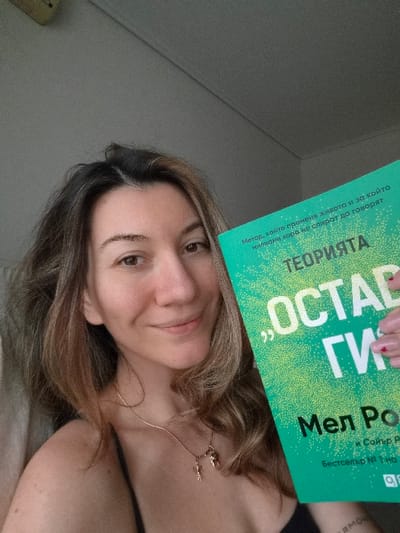 A medium-close-up image depicts a woman holding a book. The woman, appearing to be in her 20s or 30s, is positioned slightly off-center to the left, facing forward with her head slightly tilted to her right. She has light brown hair with visible darker roots, light brown eyes, and a faint closed-lip smile. Freckles are visible on her shoulders and upper chest. She wears a thin black strap or top and a thin gold necklace with two small gold charms, one resembling a stylized human figure. A partially visible tattoo beginning with "ERMO" is present on her inner left forearm. Her right hand, featuring red nail polish and a small heart design on one nail, holds a lime-green book with white text. The visible text on the book cover is in Bulgarian, including the title "ТЕОРИЯТА „ОСТАВИ ГИ“" (The Theory "Leave Them") and the author's name "Мел Роббинс" (Mel Robbins). Additional text states: "Метод, който променя живота и за който милиони хора не спират да говорят" (A method that changes lives and that millions of people don't stop talking about) and "БЕСТСЕЛЕР № 1 на" (BESTSELLER #1 on). The book's cover has a textured pattern of small white dots. The background is an indistinct, light-colored indoor wall or ceiling, featuring a visible diagonal line in the upper section indicating a corner or structural element. The specific location within the Municipal Unit of Corinth, Greece, cannot be determined from the visual information provided in the image.