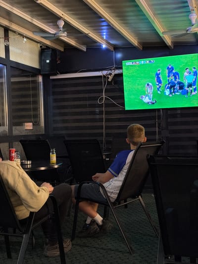 The image depicts an indoor, covered seating area, possibly an outdoor cafe or sports bar, in Nević Polje, Bosnia and Herzegovina. A large, flat-screen television mounted on the ceiling displays a live soccer match, with a scoreboard indicating 60:50 minutes played and a score of 1-0. Several individuals are seated, facing the television. In the foreground, a young boy wearing a blue and white soccer jersey is seated in a black chair. To his left, another person is seated, wearing a light-colored hoodie and dark pants. A small round table is positioned between them, holding a red soda can and a small glass bottle. The environment appears to be dimly lit, with striped blinds visible on the walls and ceiling fans overhead. The floor is covered in green carpeting. The overall scene suggests a casual gathering to watch a sporting event.