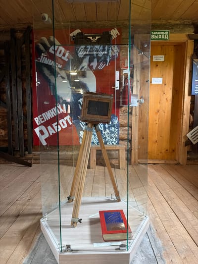 The image shows an indoor exhibit, likely a museum or historical display, featuring a vintage camera on a tripod encased in a glass display case. The camera is a wooden bellows camera with a lens. A red book with gold lettering and a blue insert is placed on the base of the display case. Behind the display case, a large red poster with white lettering and images of hands and faces is visible. To the right of the poster and display case, there is a wooden door with two signs above it, one green and one white, both reading "ЗАПАСНЫЙ ВЫХОД" (Emergency Exit). The surrounding environment consists of wooden walls, a wooden ceiling with visible beams, and a wooden floor. The overall impression is of a rustic, potentially historical setting. The location is identified as Ust'-Pyzas, Russia.